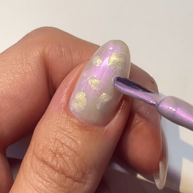 A close-up image of a hand decorating a nail with a light purple nail polish and gold-colored dots, while a brush is used to apply the nail designs.
