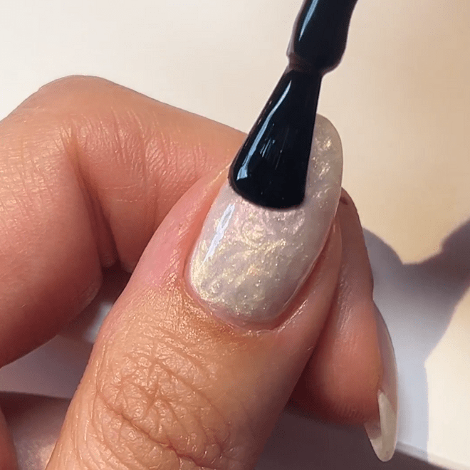 Female hand with a pearlescent nail, while a brush applies nail polish or coating.