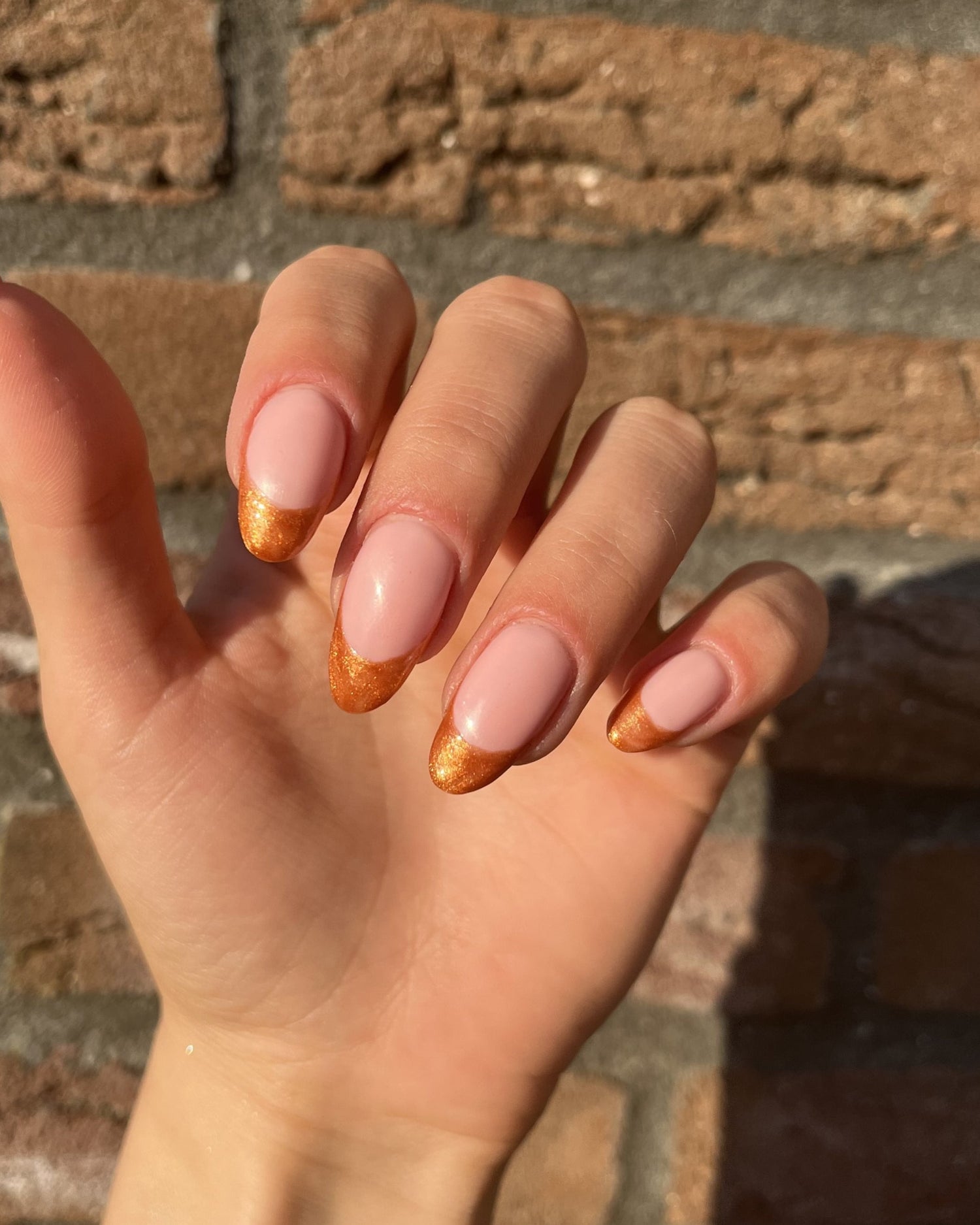 A hand with nails in a French manicure style, with pastel pink base and golden shimmering ends, against a stone background.
