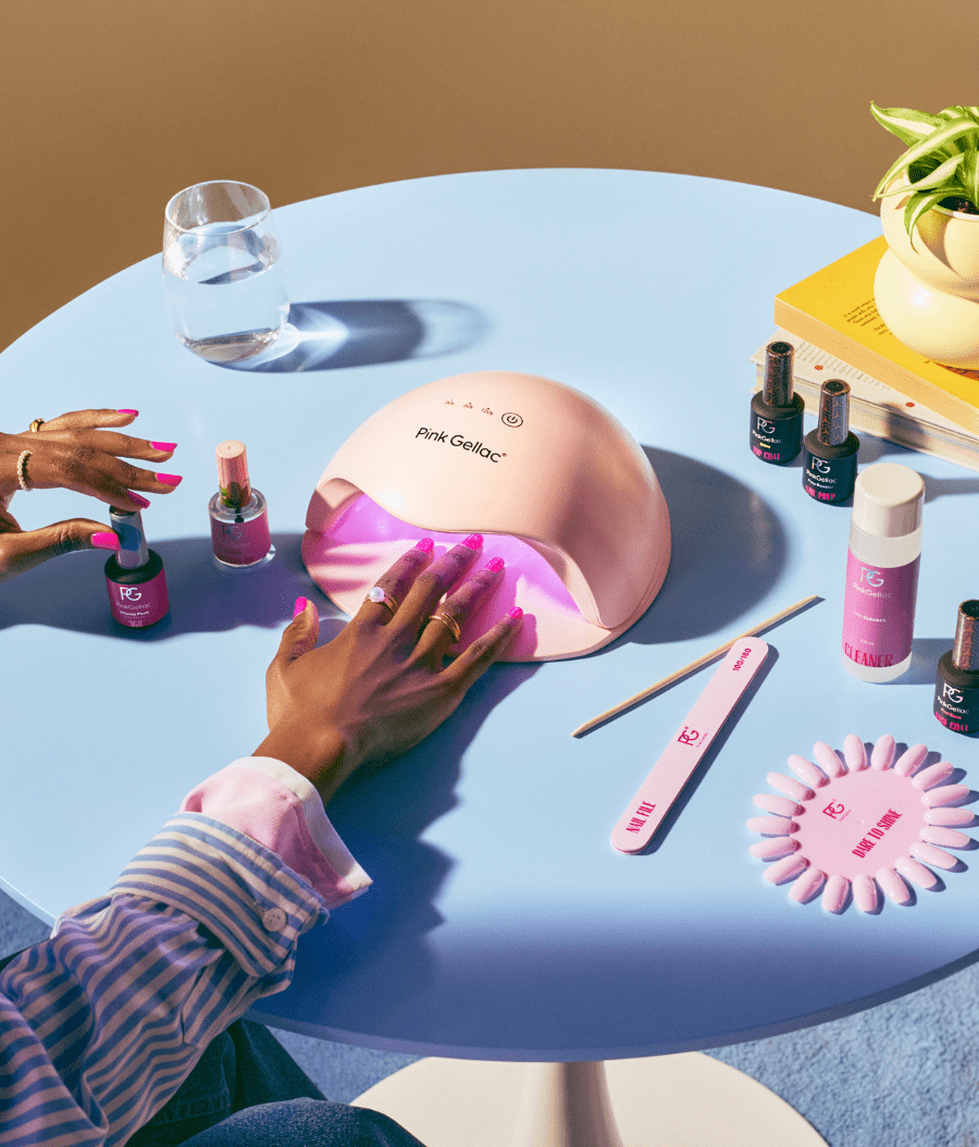 Person uses a pink UV lamp to dry nail polish on a blue table, surrounded by nail care products such as nail polish bottles, a nail file, and a glass of water.