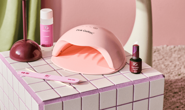 A setup of a pink nail dryer, nail polish, a file and a bottle of liquid, placed on a white and purple tiled cube, with a pink and green background.