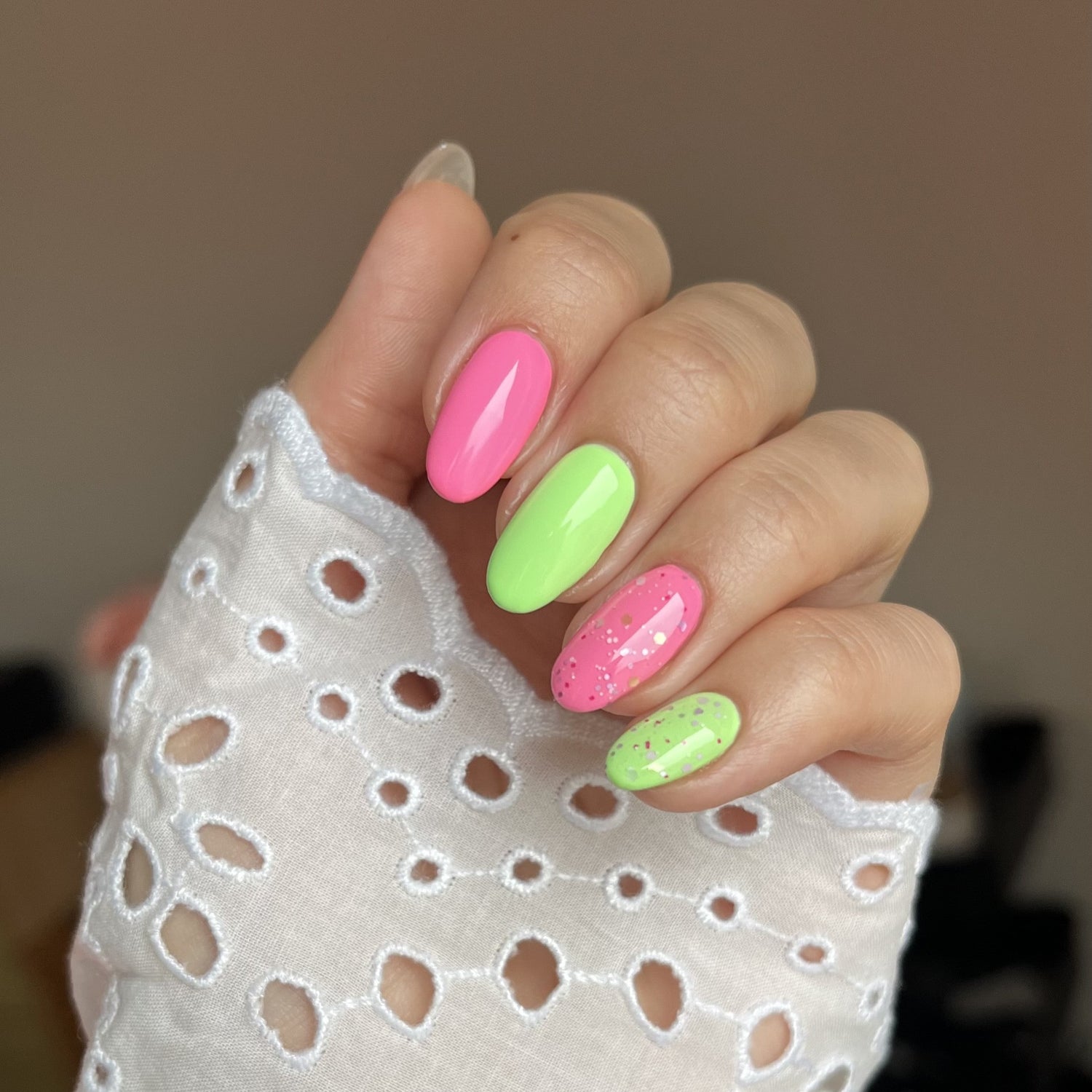 A hand with bright pink and bright green nail polish and glitter, against a white lace fabric with a hole pattern.