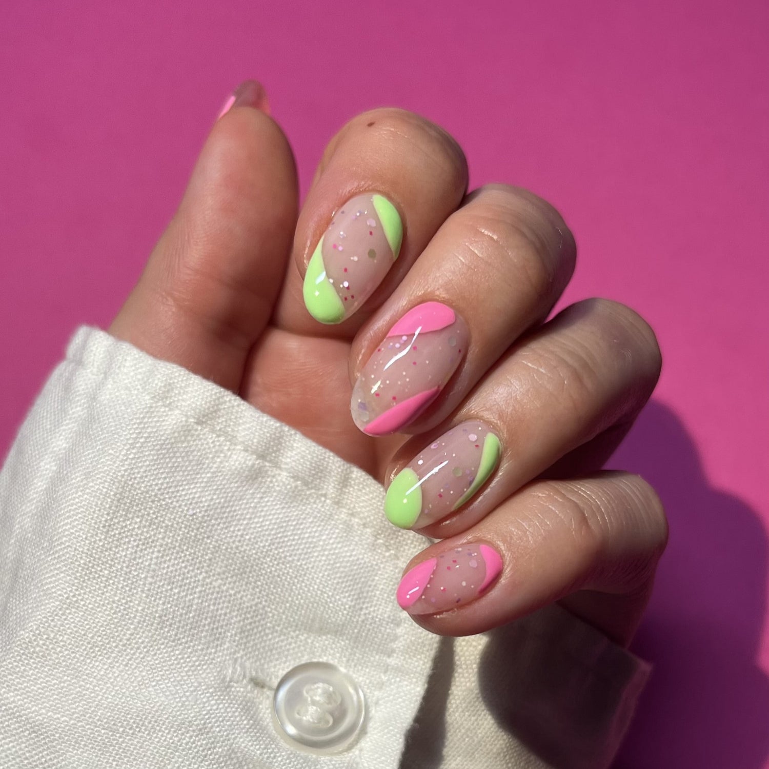 Close-up of a hand with colorful nail art in neon pink and green with glitter details, against a pink background.