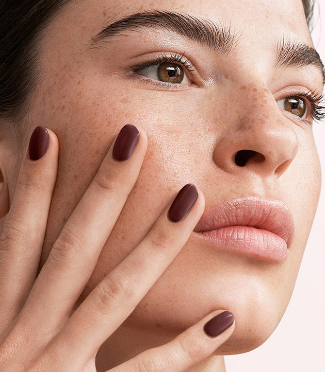 Close-up of a freckled woman touching her face with her hand, wearing dark red nail polish.