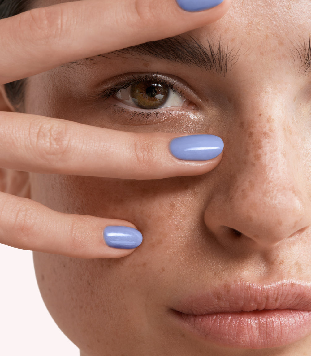 Close-up of a face with freckles, holding fingers with lilac painted nails around one eye.