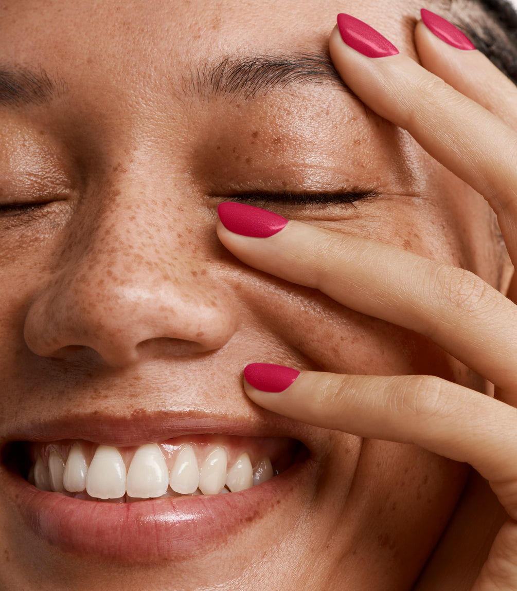 Close up view of a smiling person with closed eyes and spread fingers with pink nail polish on their face.