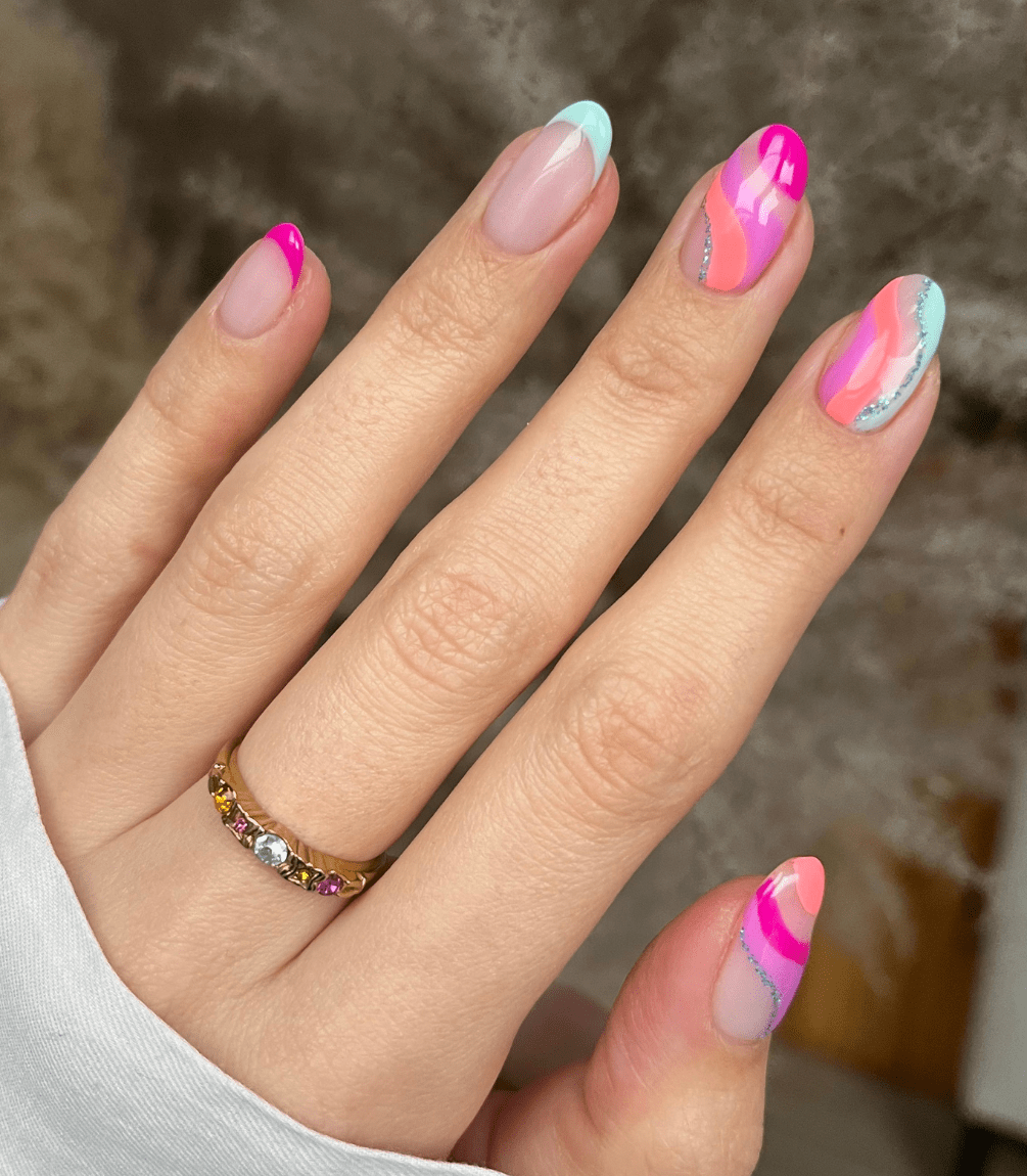Hand holding brightly colored and artistically designed nails, including pink and blue tones, with a gold ring adorned with colored stones on the ring finger.