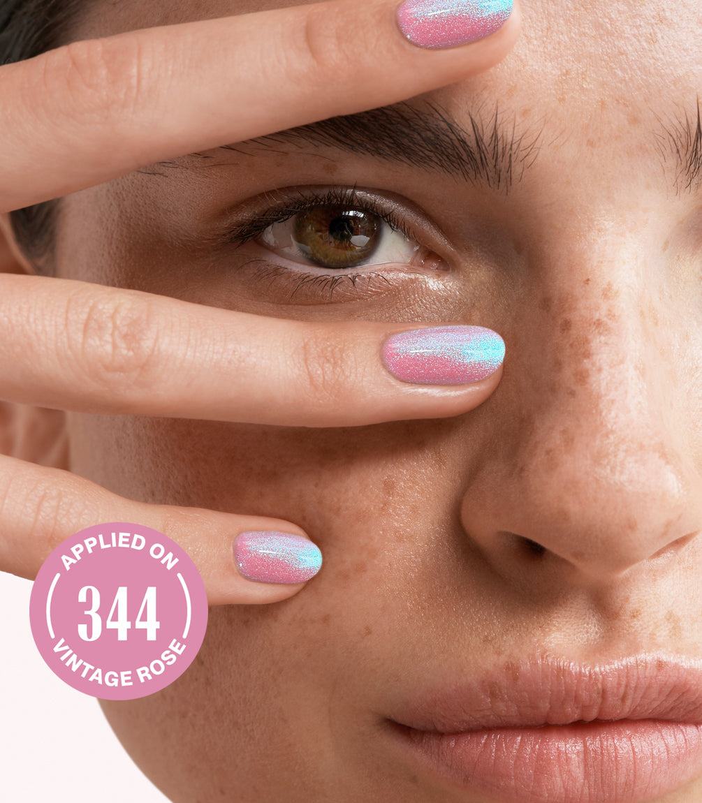 Close-up of a freckled face, with fingers with shimmering pink nail polish placed along an eye, and a pink badge that reads "applied on 344 vintage rose".