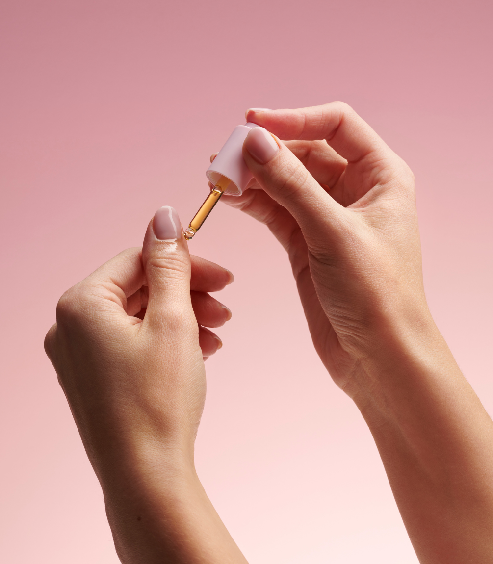 A hand applying cuticle oil to the nails with a dropper against a pink background.