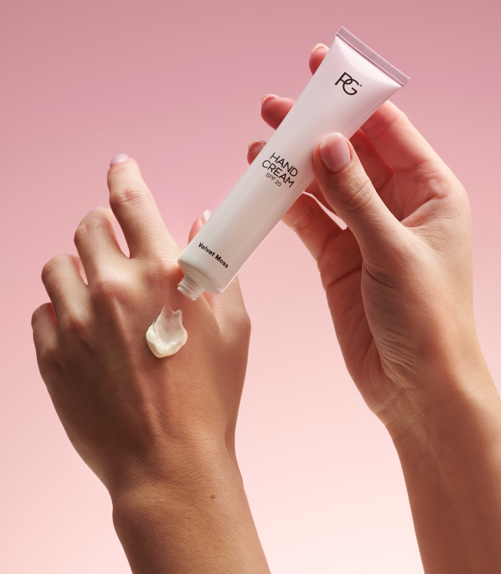 Two hands with one hand applying cream to the other hand from a tube labeled "Hand Cream SPF 20" and "Velvet More", against a pink background.