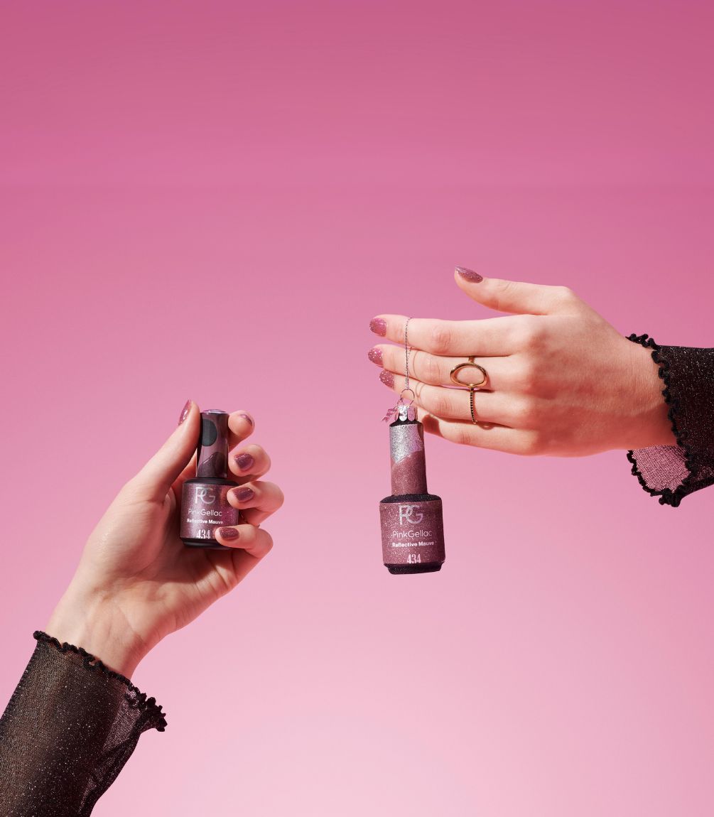 Two hands hold nail polish bottles against a pink background.