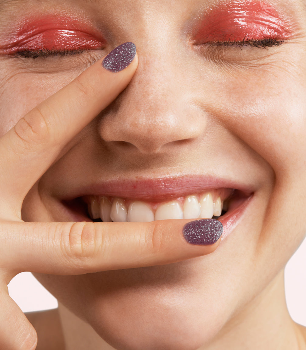 A smiling person with bright pink eyeshadow and shiny nail polish on the fingers that lie near the mouth.
