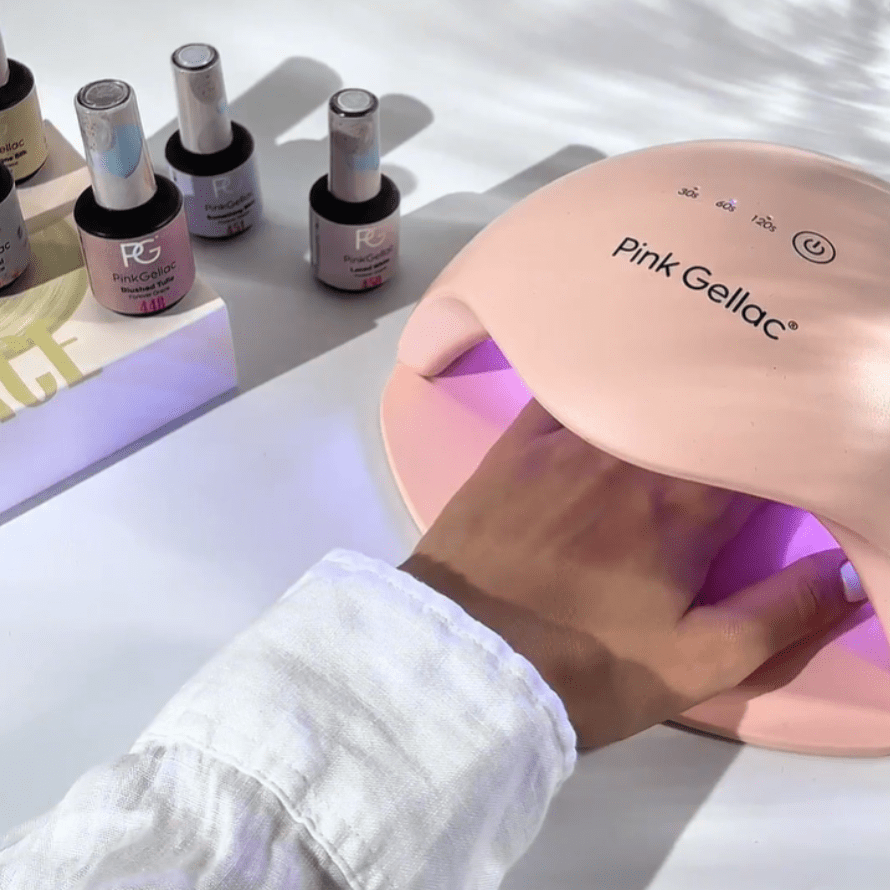 Hand under UV lamp for nails, with Pink Gellac bottles and packaging on a table.