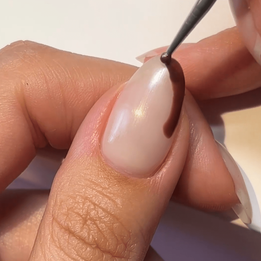 Person applies brown nail polish to an almond-shaped nail with a thin brush.
