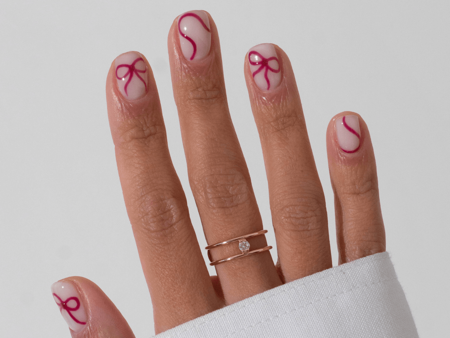A hand with pink ribbon-like nail art and a gold ring with a small diamond on the ring finger.