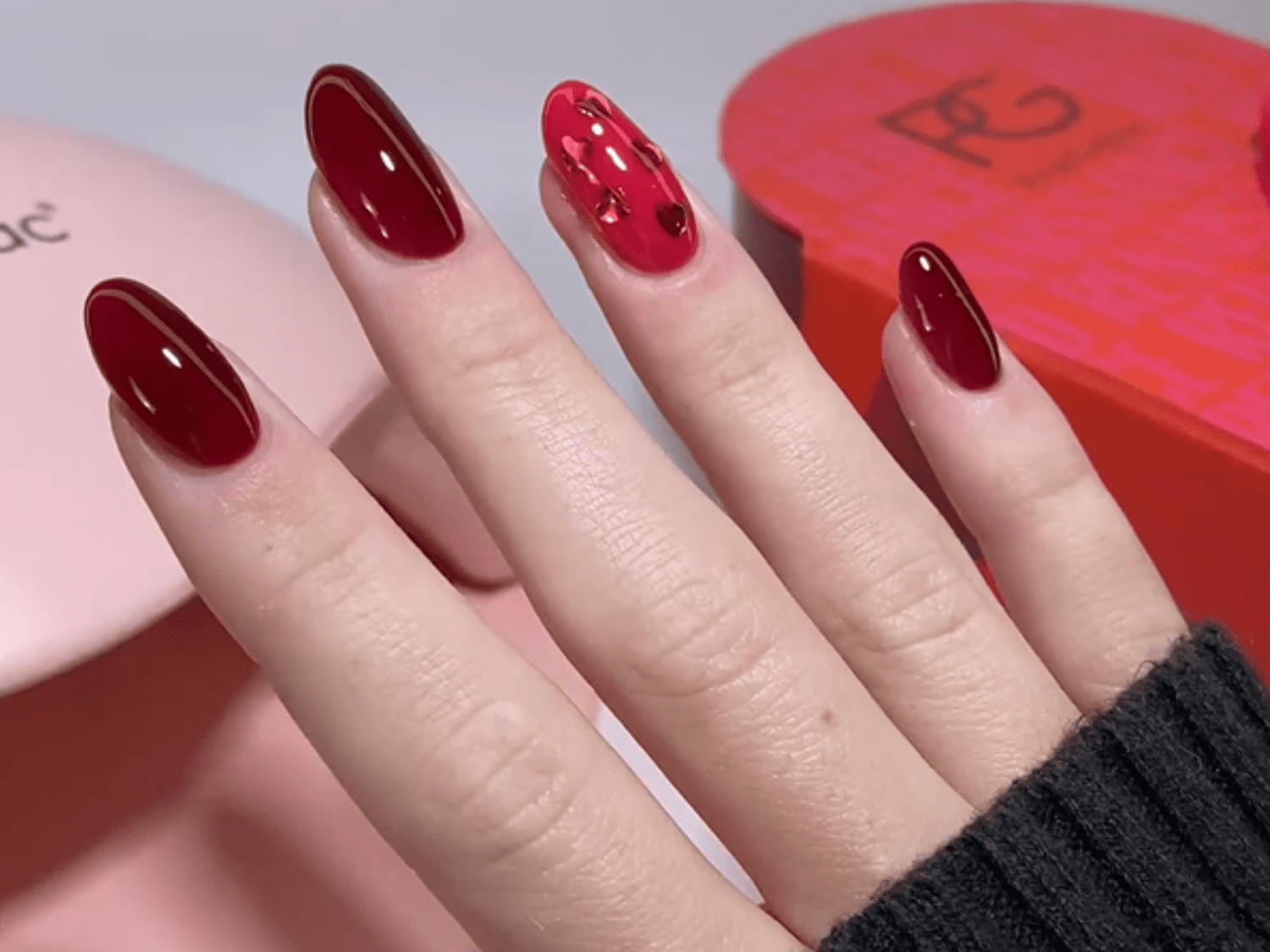 Close-up of a hand with shiny dark red nail polish, with the ring finger having a pink tint with small hearts.