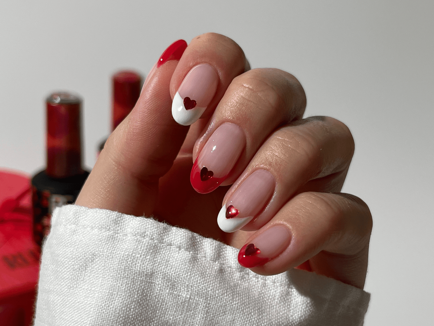 A hand with nails in a French manicure style, decorated with red and white hearts, against a background with blurry nail polish bottles.