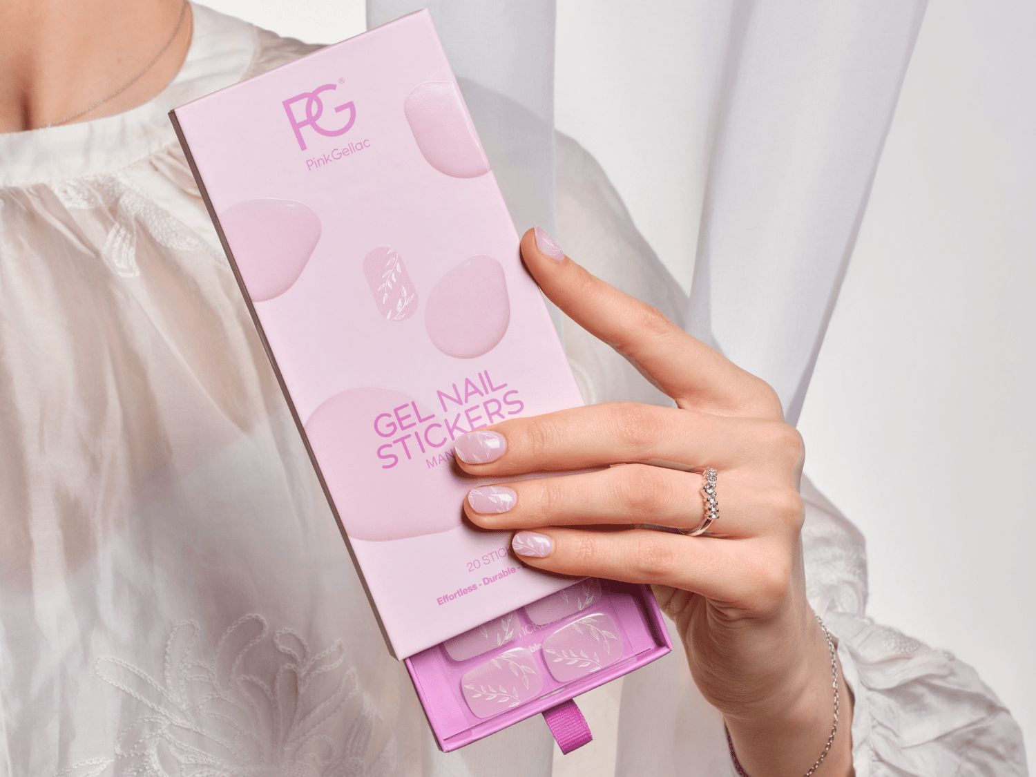 Woman with gel polish stickers holding pink packaging, well-groomed nails, white lace blouse visible.