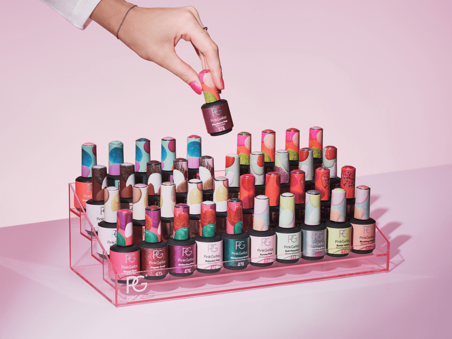 Hand takes a bottle of nail polish from a rack of colorful bottles against a pink background.