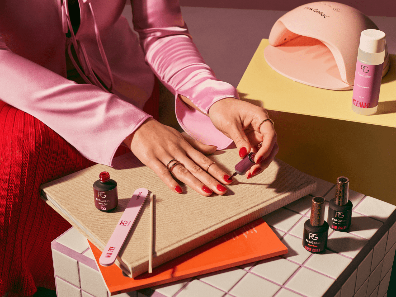 Person painting nails with pink nail polish, surrounded by manicure supplies and a nail UV lamp.