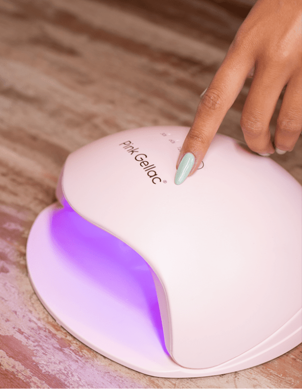 Female hand with green nail polish presses the on button of a pink Pink Gellac nail dryer with purple light.