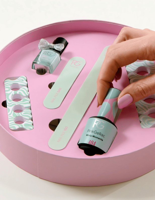 A round, pink box of manicure accessories, including nail polish in a blue shade, nail files, and a finger spacer; One hand takes the nail polish bottle.