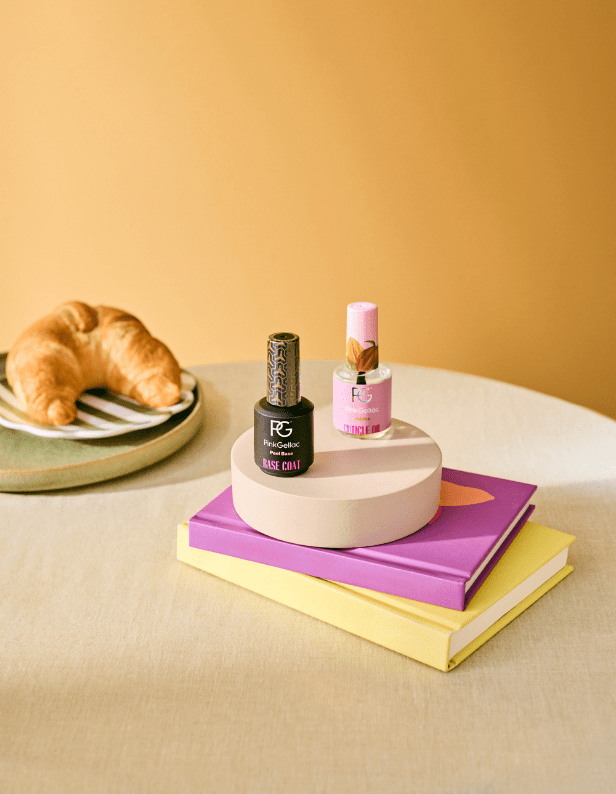 Two bottles of nail polish on a round stand, placed on top of a stack of books with a croissant in the background against a yellow background.