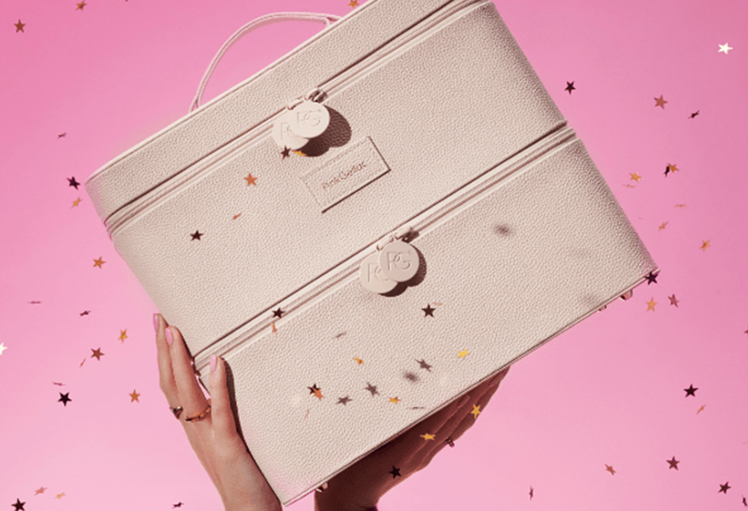 A hand holds a pink suitcase against a pink background with star confetti.