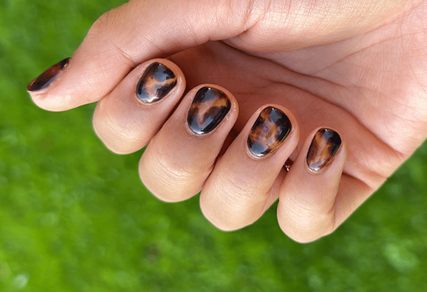 Hand with short painted nails in tortoiseshell pattern, against a blurred green background.