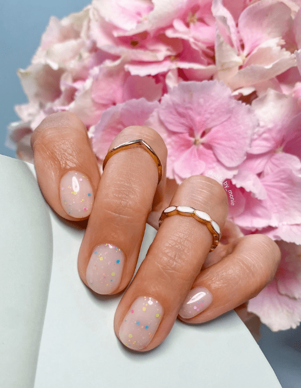 Hand holding pastel pink nails with colored dots, ornate rings, and pink hydrangeas in the background.