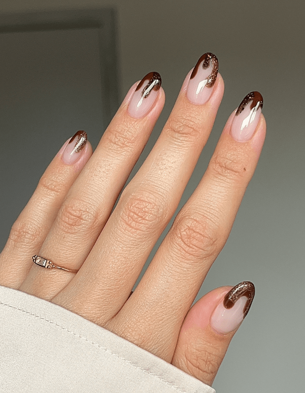 A hand with almond-shaped nails, painted with a shiny brown and gold drop pattern on a neutral base, and a thin ring on the ring finger.