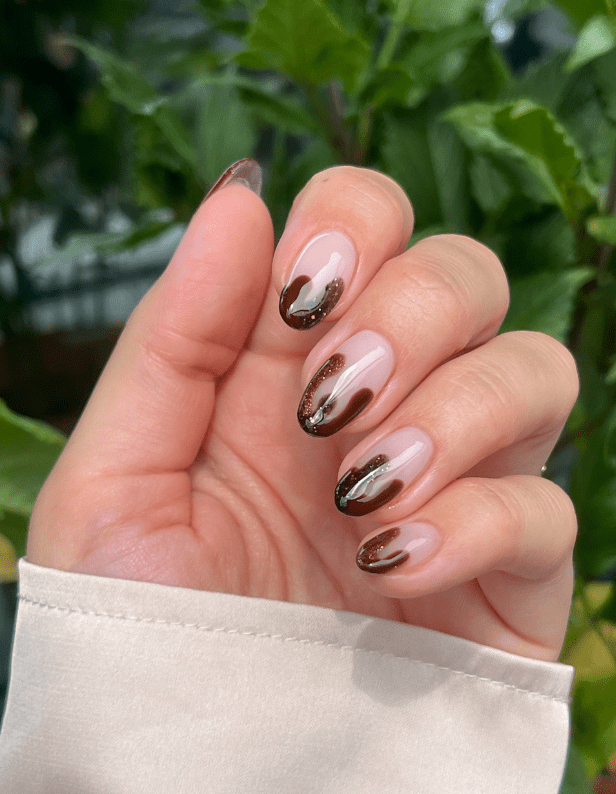A hand with a beige shirt poses in front of green leaves, with the nails painted with a shiny brown and black wave pattern.
