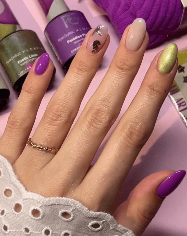 Hand holding colorful nails, including purple, pink with silver trims, nude and green nail polish, against a background with nail polish bottles.