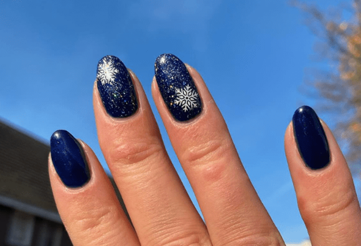 A hand with dark blue nail polish with snowflake patterns on two nails against a blue sky.