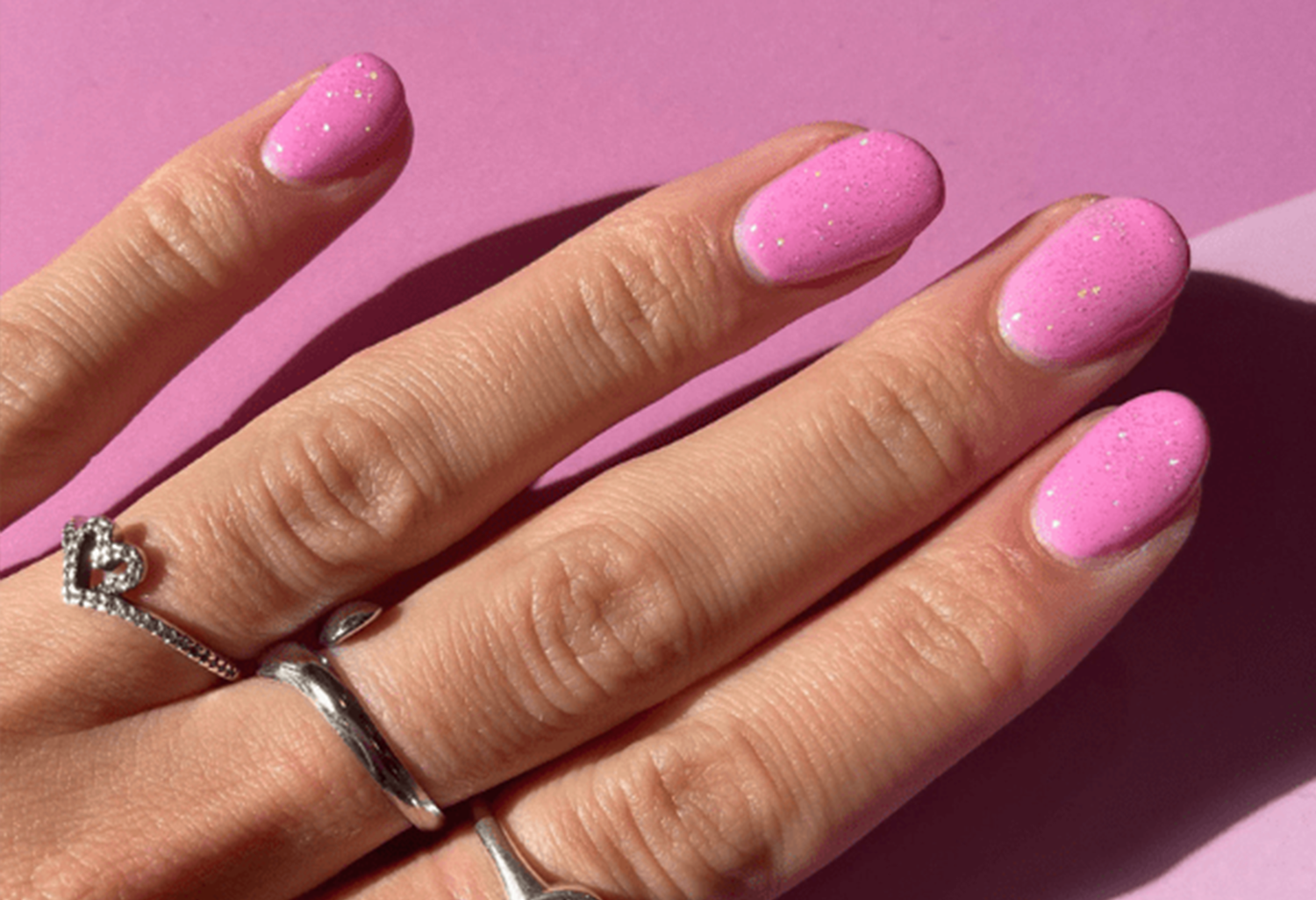 Hand holding pink glitter manicure on a pink background, with silver rings on the fingers.
