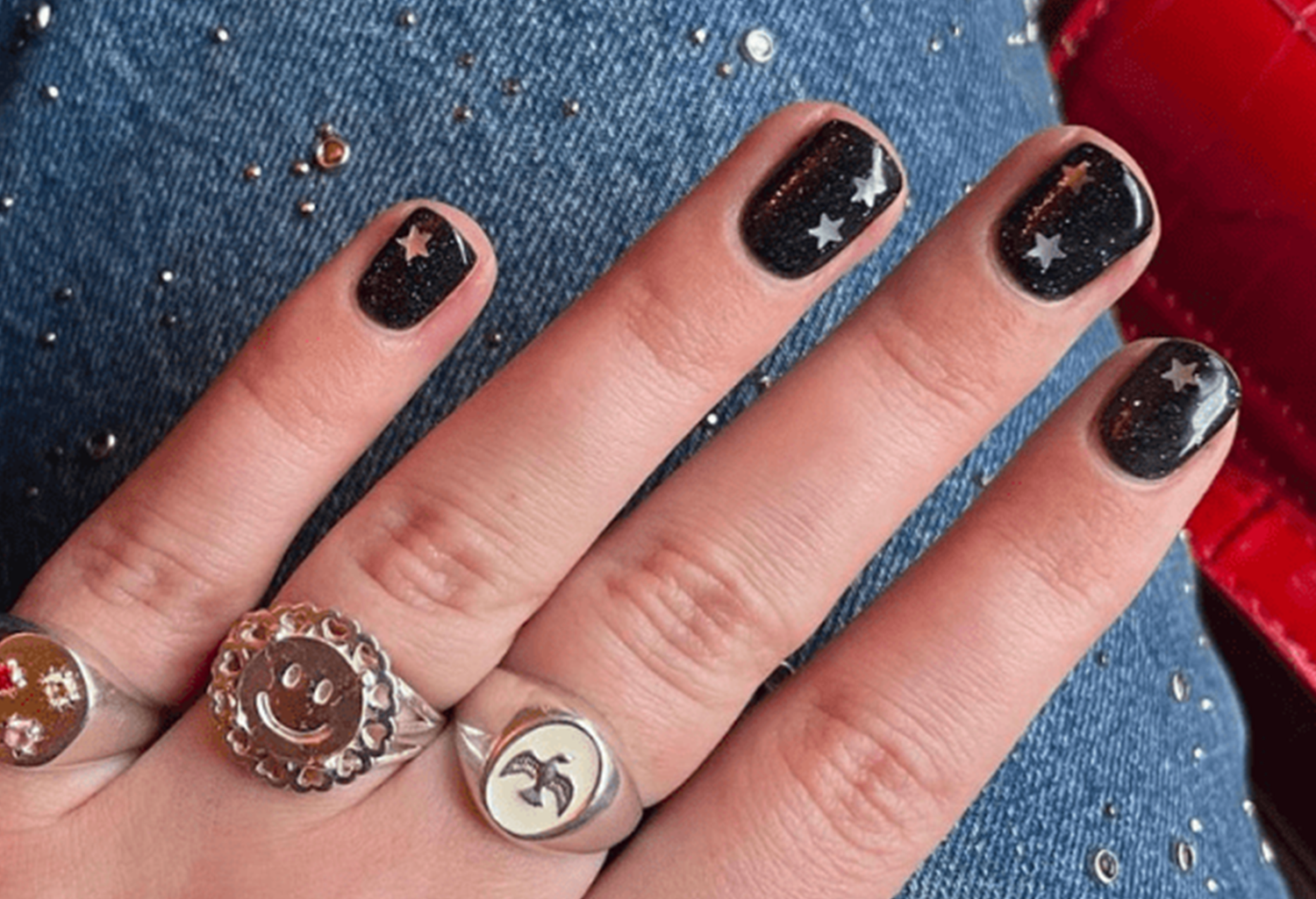 Hand with black nail polish and stars, silver rings with different designs, on jeans.