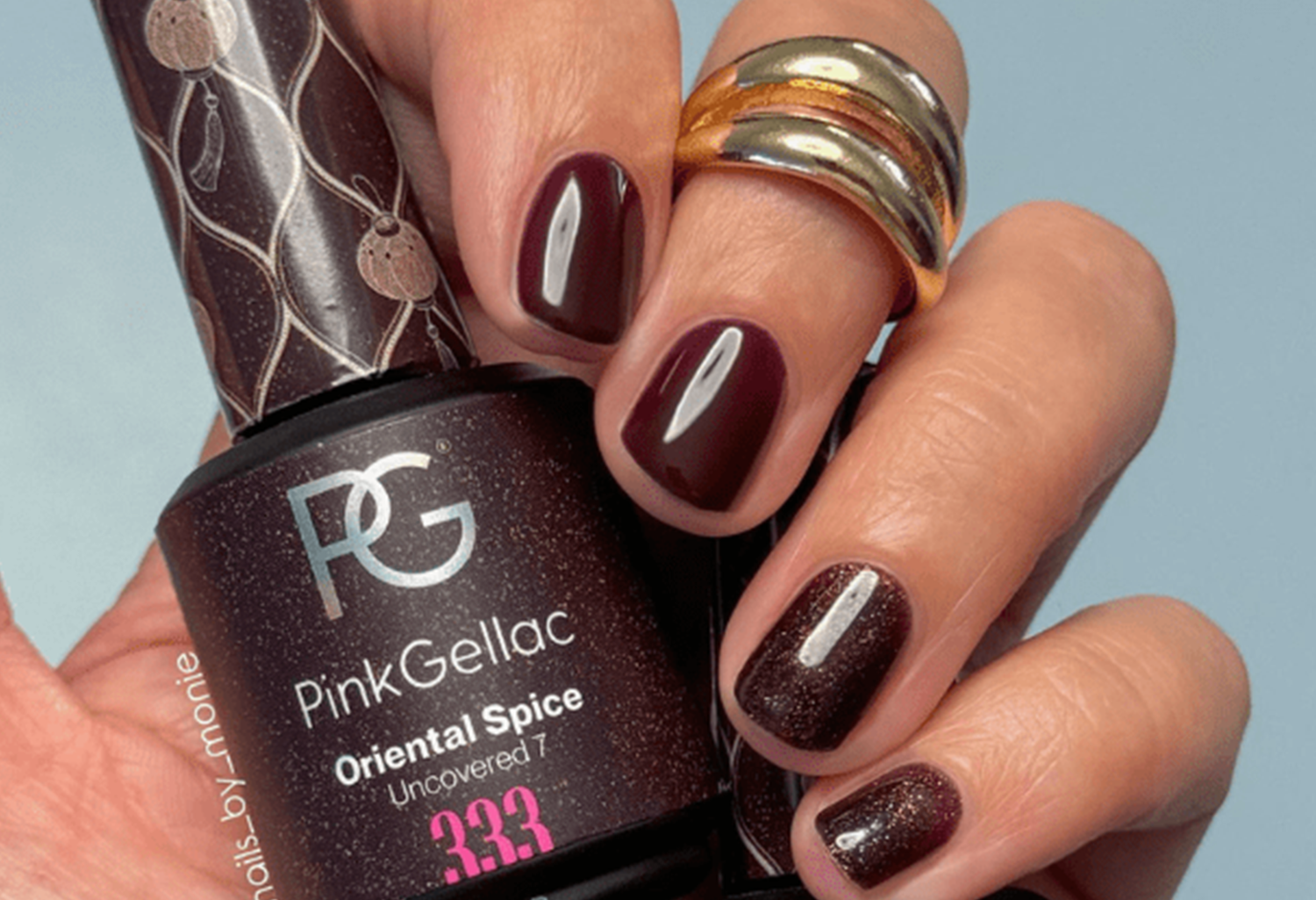 Close-up of a hand holding burgundy nail polish and a gold-colored ring, while the person holds a bottle of Pink Gellac with the color "Oriental Spice".
