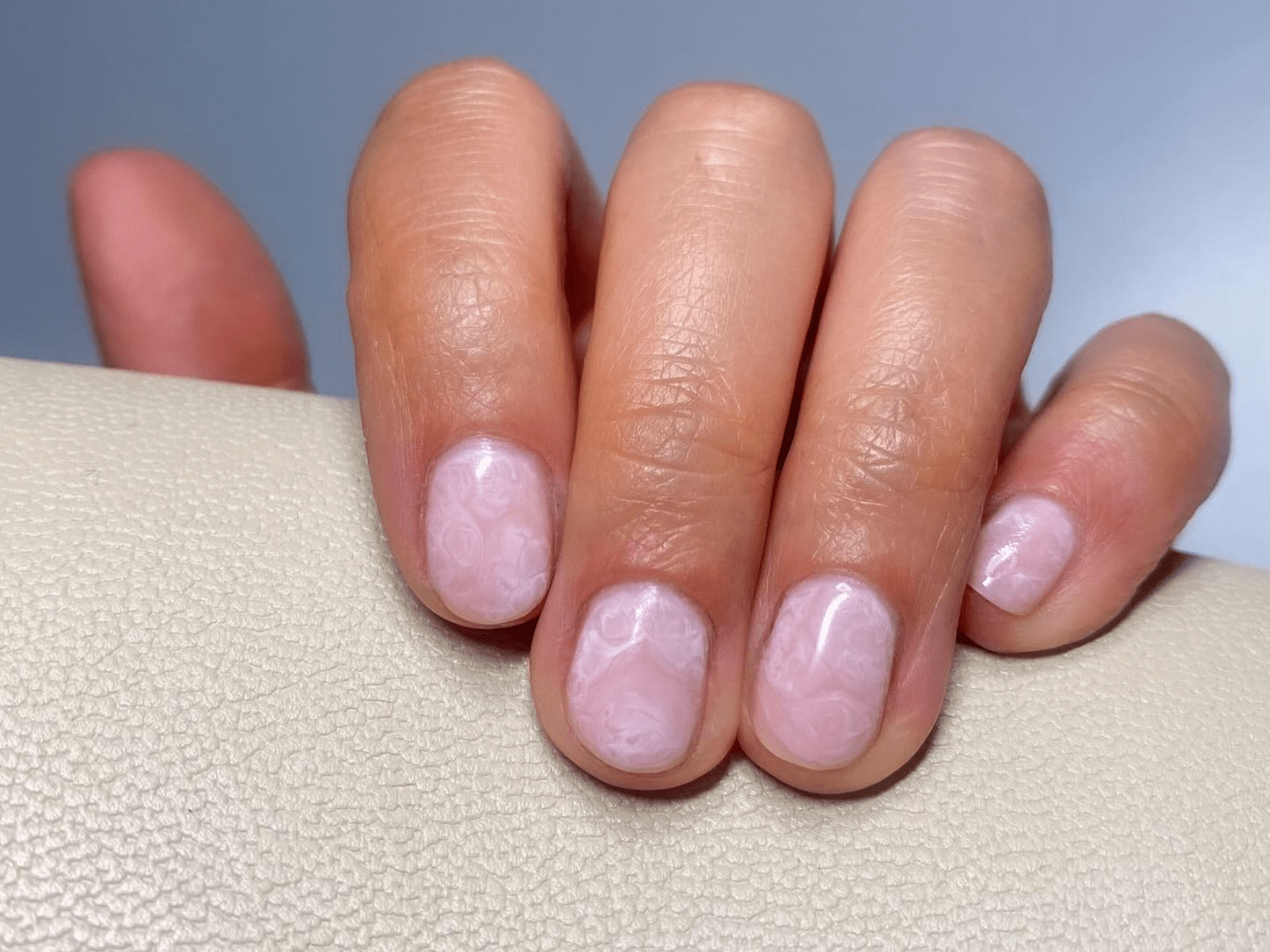 Hand with short nails, painted in a light pink color with a marble pattern, resting on a lightly textured background.