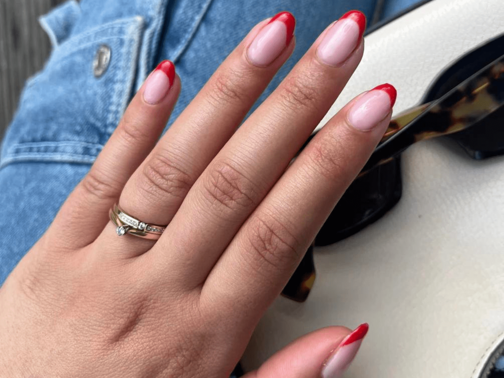 A hand with red-pink manicured nails wears a ring, against a background of a cream-colored bag and jeans.