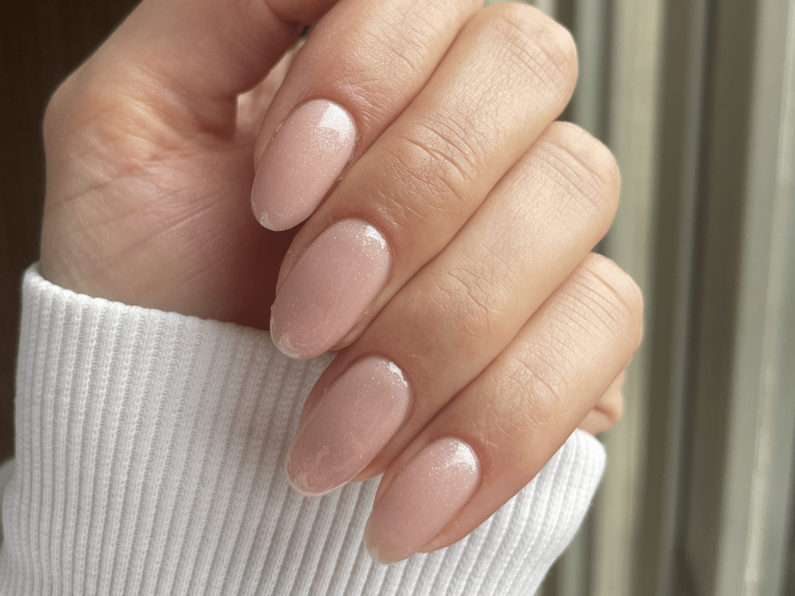 Close-up of a hand with pinkish-yellowish, shiny, almond-shaped nails, wearing a sweater with white sleeves.