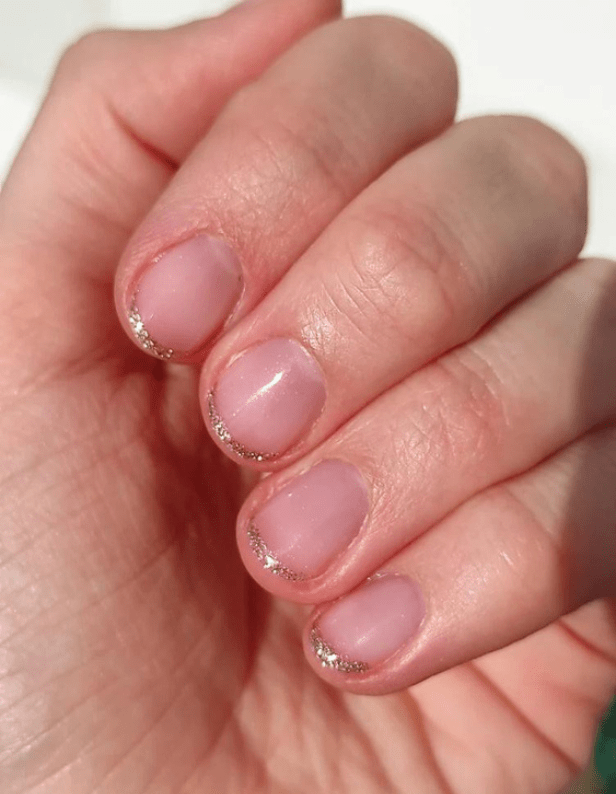 A hand with short-cut nails with a subtle pink shine and silver glitter at the nail edge.