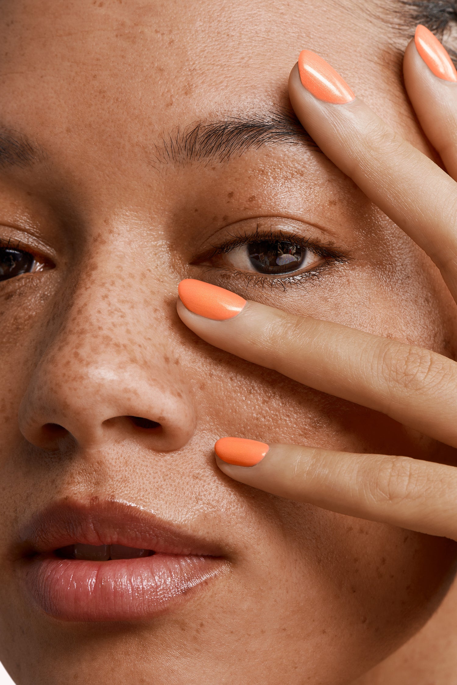Close-up of a face with freckles and orange nail polish on the fingers resting against the skin.
