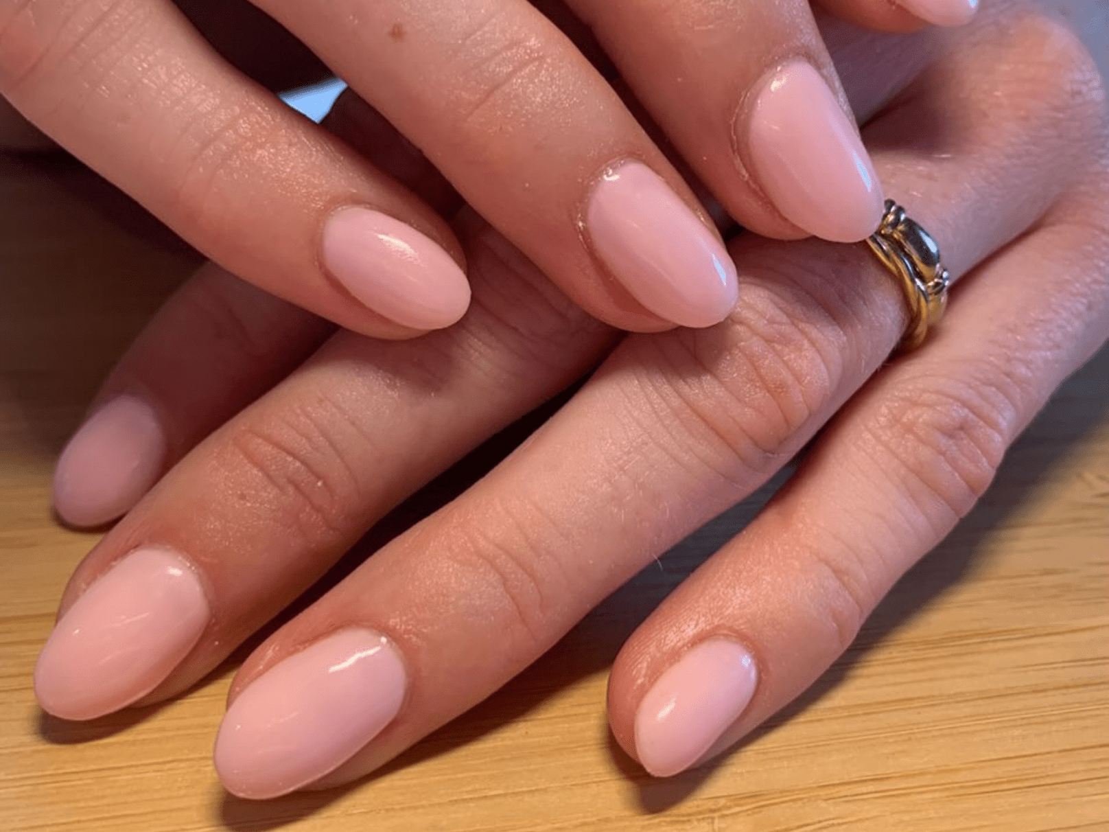 Close-up of two hands with pink painted nails and a gold ring, lying on a wooden surface.