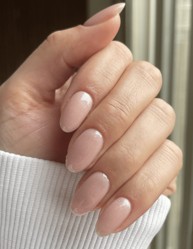 A hand with long, almond-shaped nails, painted in a subtle, light pink color with a glossy finish, against a neutral background.