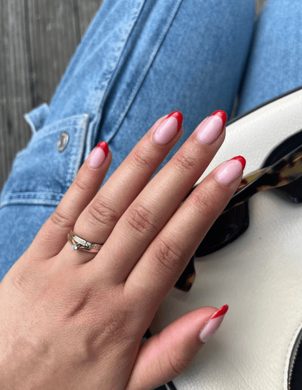 A hand with long nails painted pink with red tips wears a ring with a diamond, resting on a lap with blue jeans and a white bag.