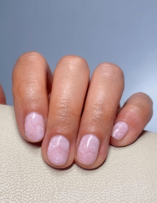 Hand with short-cut nails painted in a soft pink shade with subtle marble pattern, resting on a light-colored background.