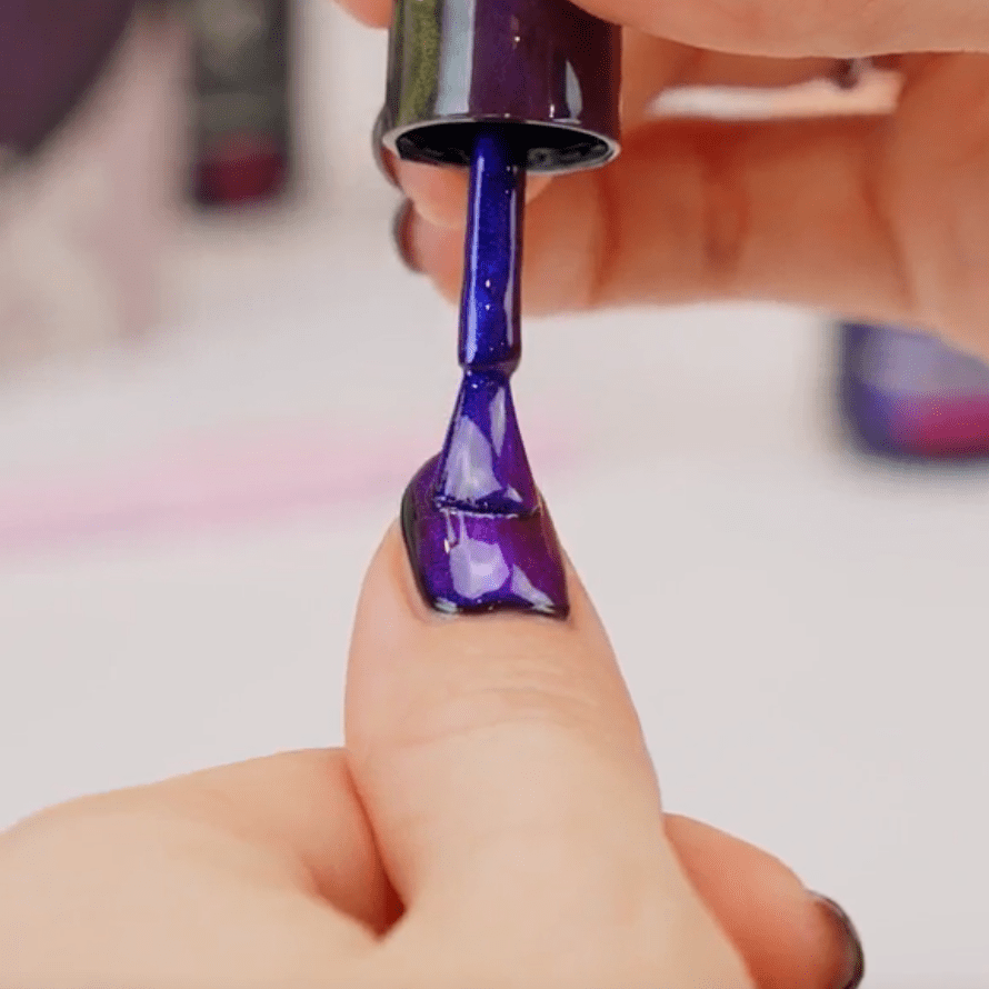 A thumb on which purple nail polish is applied with a brush from a bottle.