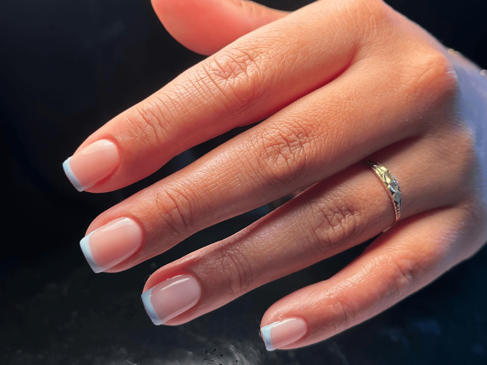 A hand with a gold ring and a French manicure on black background.