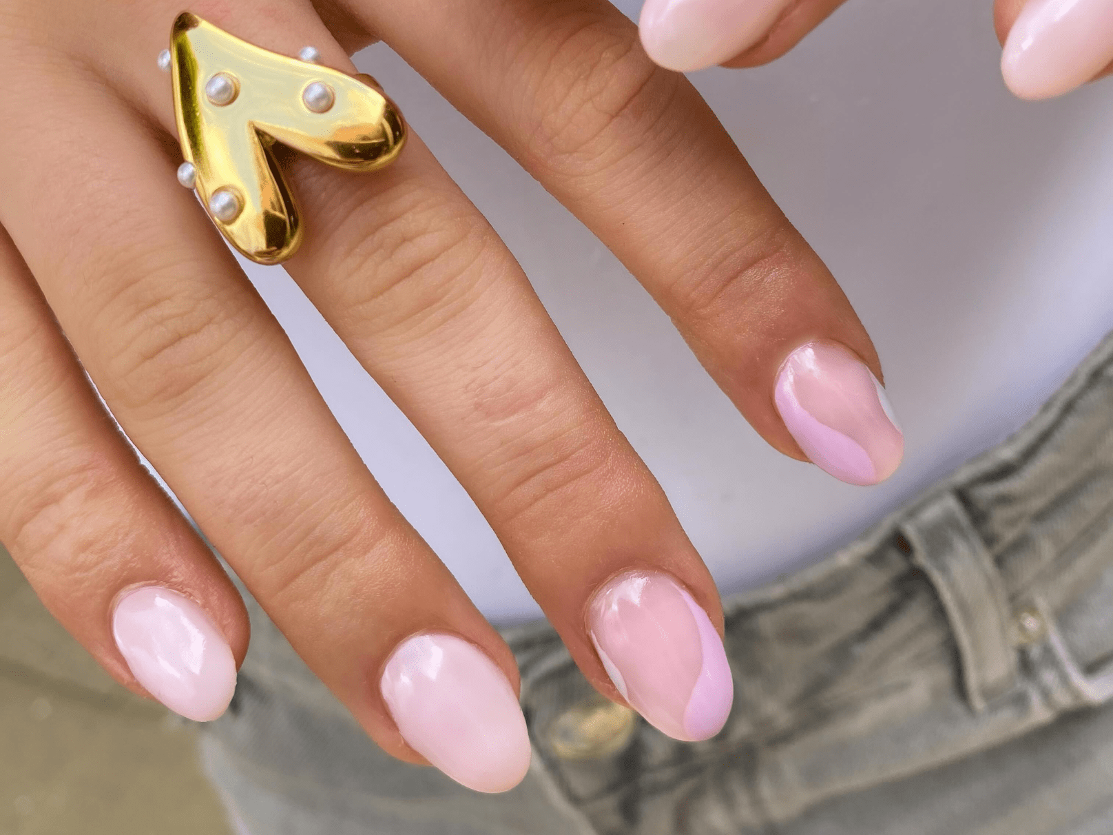 Close-up of a hand with light pink painted nails, a striking gold-colored ring with pearls in the shape of a playful arrow and pants in a light color in the background.