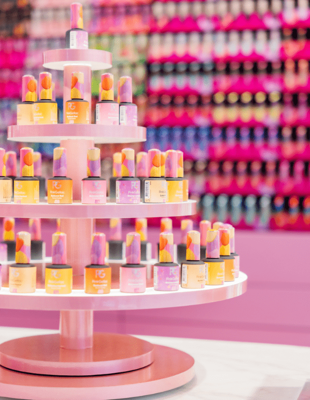 Round display full of brightly colored nail polish bottles in a store, with a background of pink and colorful products.